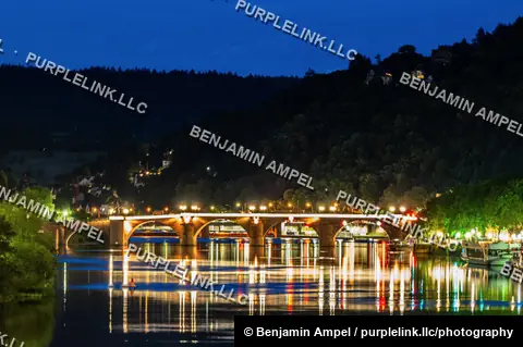 The stone arches of Heidelberg's Old Bridge glow with lights and reflect on the Neckar river beneath a wooded hillside at dusk.
