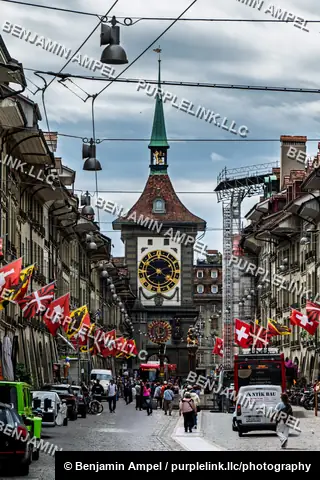 Bern, Switzerland - June 2026: The Zytglogge clock tower stands at the end of a bustling street decorated with Swiss flags and lined with shops and vehicles.