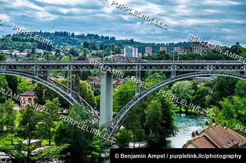 A steel arch bridge crosses the turquoise Aare river amid green parkland and rooftops on the edge of Bern.