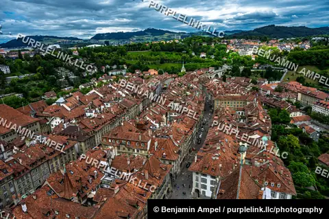 Rows of red-tiled rooftops fill Bern's historic old town, framed by green hills and a distant bridge under a cloudy sky.