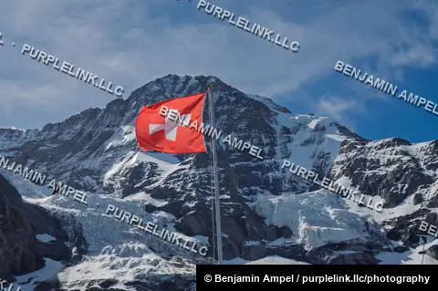 A Swiss flag flies in the wind against a backdrop of a snow-covered alpine peak and glacier.
