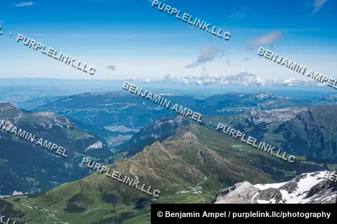 Rolling green alpine ridges descend toward a distant valley town seen from high above at Jungfraujoch.