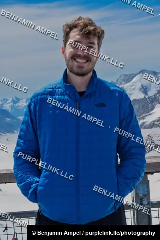 Jungfraujoch, Switzerland - June 2026: A smiling man in a blue jacket poses on a viewing platform with snow-covered mountains behind him.