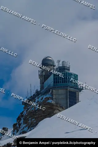 The Sphinx Observatory research station sits atop a rocky snow-covered summit at Jungfraujoch beneath a cloudy sky.