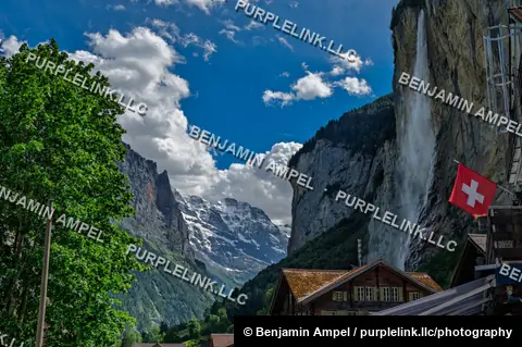 A tall waterfall cascades down sheer cliffs beside wooden chalets in the Lauterbrunnen valley, with a Swiss flag flying and snow peaks visible beyond.