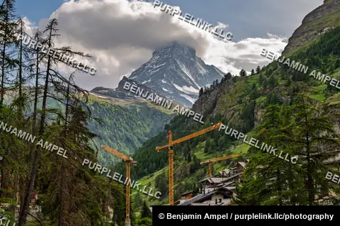 Zermatt, Switzerland - June 2026: The Matterhorn emerges from clouds above wooden chalets, a building marked Magnolia, and construction cranes.
