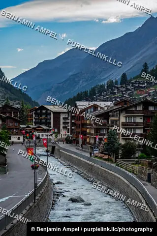 Zermatt, Switzerland - June 2026: A rushing mountain stream cuts through the village of Zermatt, lined with wooden chalets, shop signs, and heraldic flags.