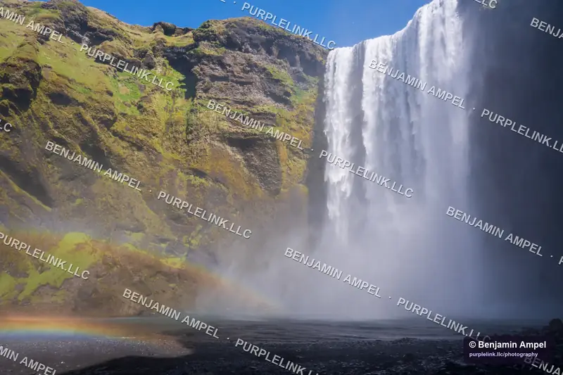 Skógafoss waterfall falling from a green cliff, with a rainbow in the spray at its base